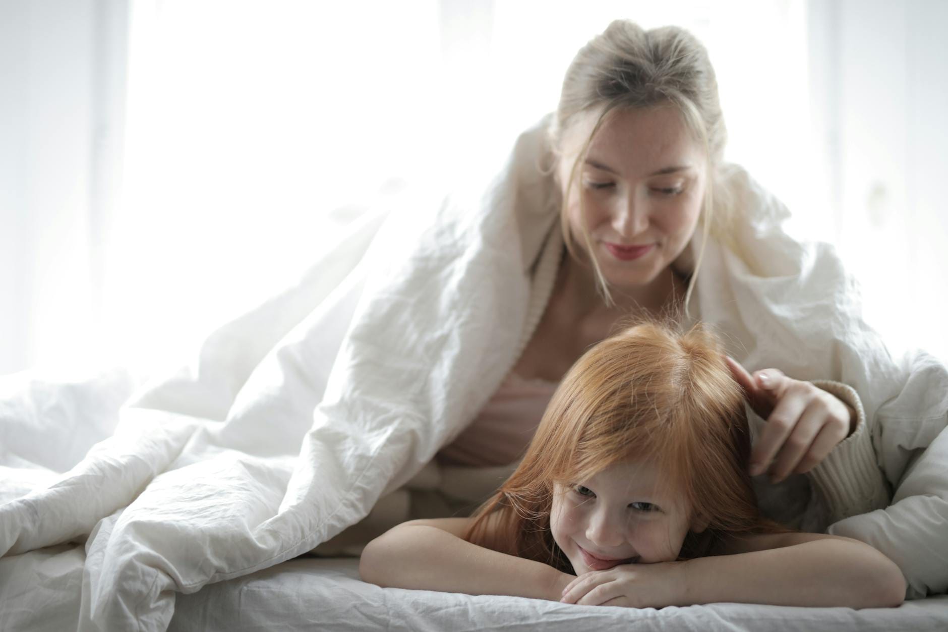 Mama przytula córkę w łóżku, razem leżą pod ciepłym kocem