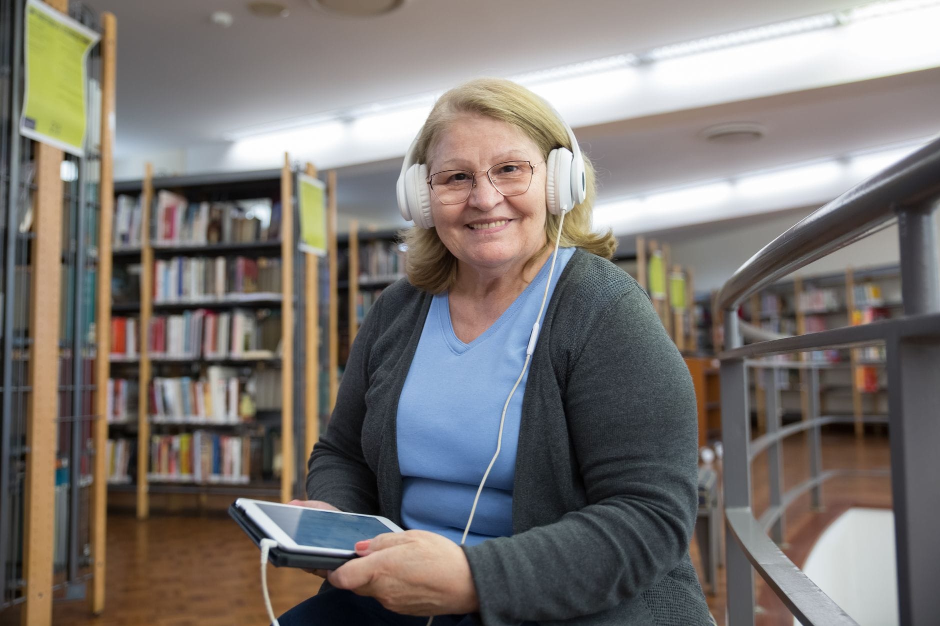 Starsza kobieta w bibliotece słucha audiobooka na tablecie